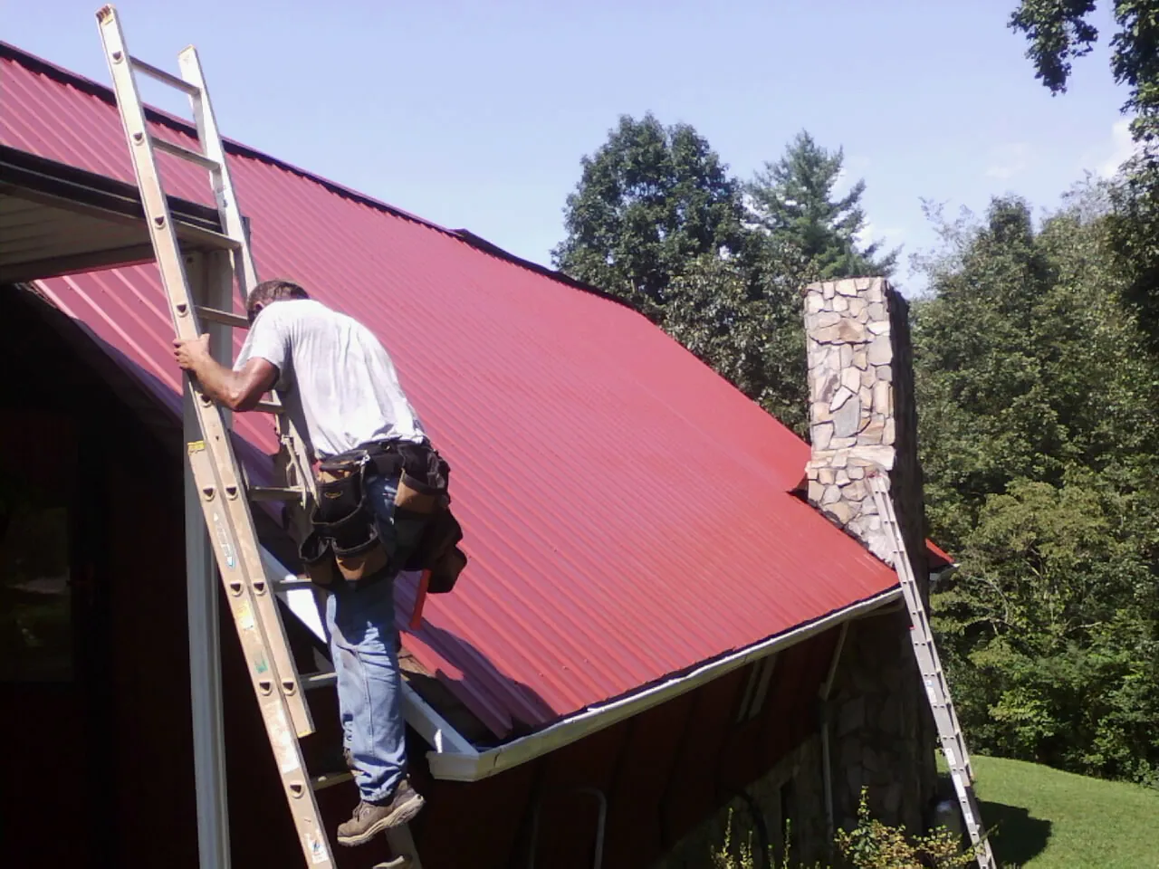 Roofer on a ladder beside a red metal roof and stone chimney — Soffit & Fascia Repair by Roofing Brothers in Oakley